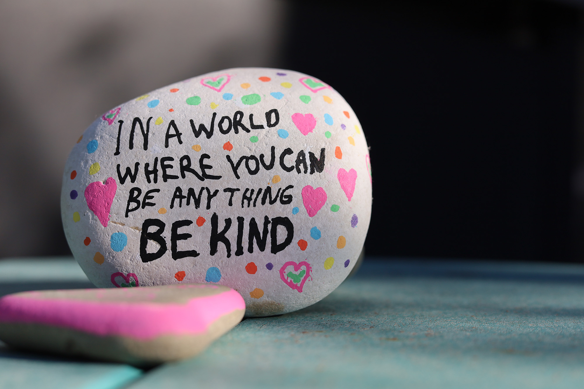 A painted rock reads "In a word where you can be anything be kind."